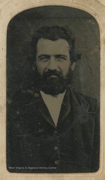 Black and white image of a white man with thick, wavy dark hair and a medium length beard looks contended into the camera. He wears a white shirt and a velvet jacket