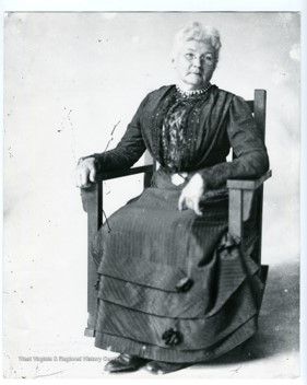 An older woman with white hair and glasses sits in a wooden chair with her arms resting on the arms of the chair. She wears a dark colored dress with layers of fabric on the skirt. She looks thoughtfully into the camera