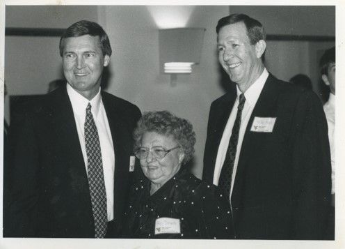Two men stand on either side of the same woman, they are all slightly younger. The men wear suits and the woman has short curly hair and glasses. They are all smiling 