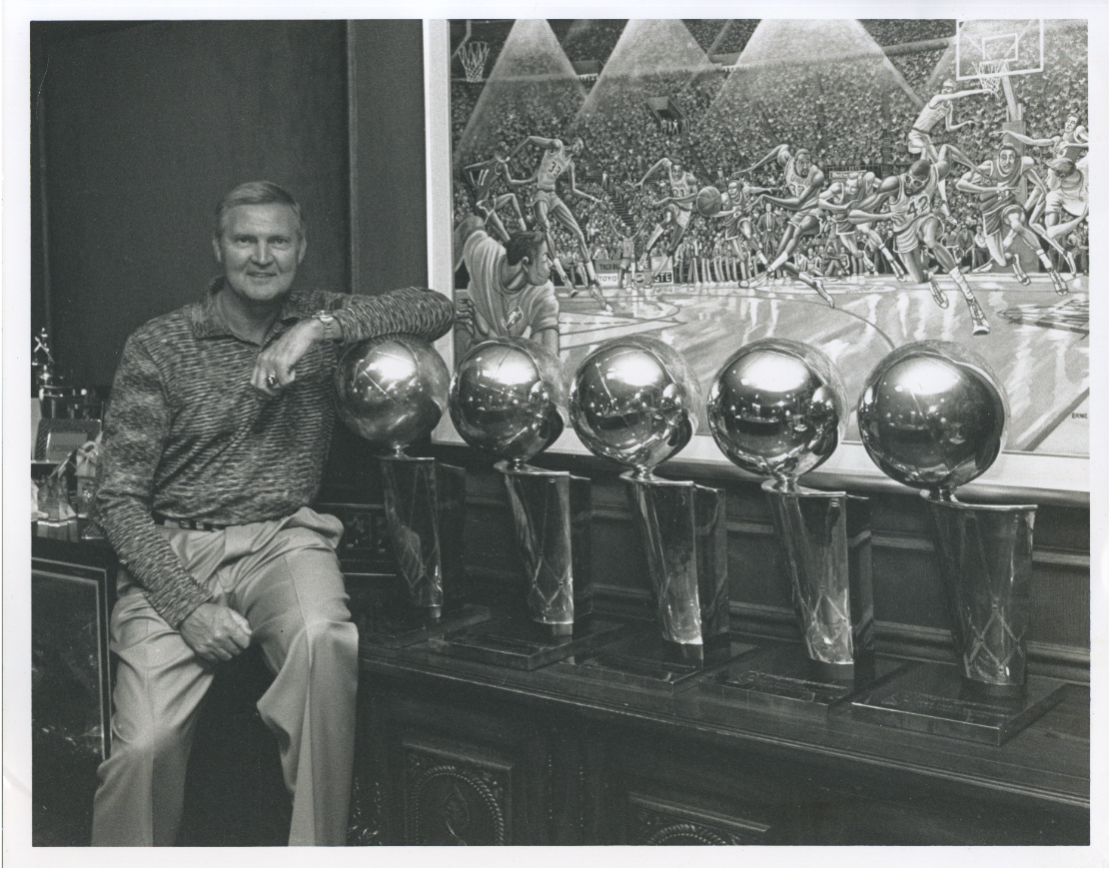 jerry sits next to five identical trophies, depicting a large ball on the edge of a hoop, leaning his elbow on the one closest to him. he is smiling. 