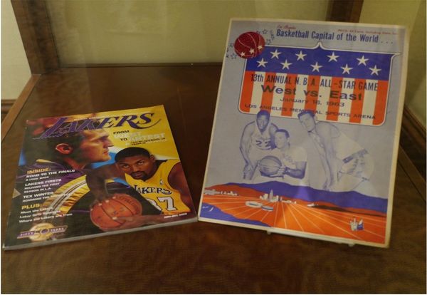 A magazine reading "Lakers" with Jerry's picture as well as another player, an african american man holding a ball. To the right is a poster advertising the east-vs-west all star game