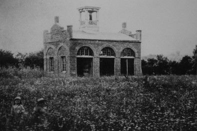 A stone engine house stands in a flat field, surrounded at the edges by trees. 